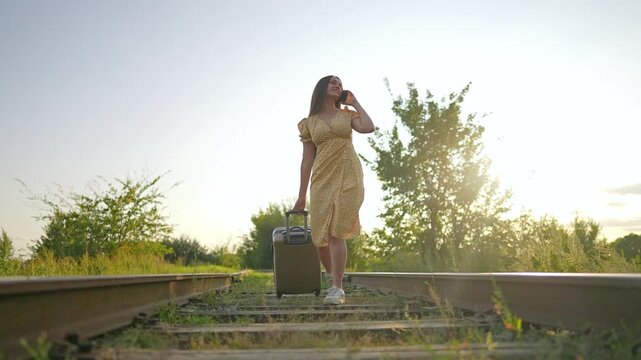 Woman stands with suitcase on abandoned railway tracks at sunset. Female traveler looks up while holding luggage on overgrown railroad. Journey pauses on rail track between trees in golden light.