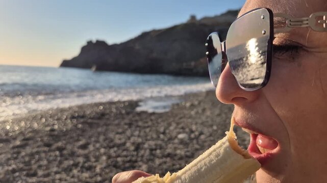 Woman eating banana on rocky beach during sunset, enjoying summer vacation. Close-up handheld shot.