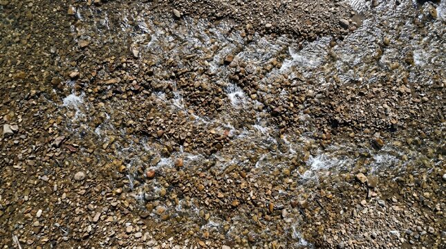 Aerial view of transparent river water rushing over textured pebbles and smooth stones in a natural stream bed landscape.
