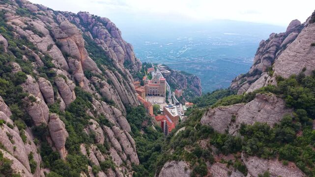 Aerial view of Santa Maria de Montserrat monastery complex tucked between dramatic conglomerate rock formations in the mountains of Catalonia, Spain, with valley below