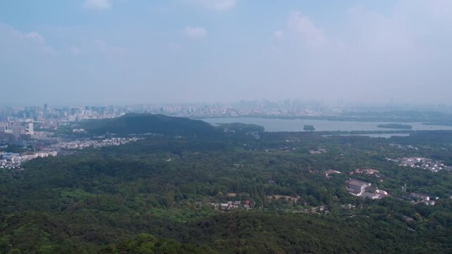 A high-angle panoramic view shows dense forest, a lake, and a distant hazy urban skyline under clear sky.