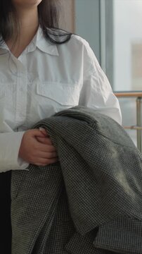 Close up of woman holding and adjusting gray houndstooth coat. Focus shifts to detail her hands gripping fabric emphasizing. Scene is set indoors by metal railings and glass walls