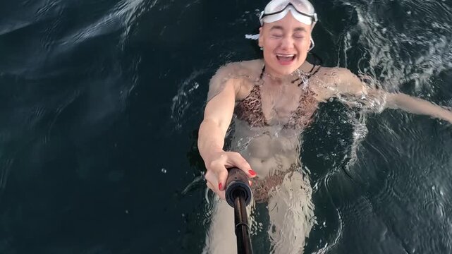 Bathing girl filming herself with monopod while floating in open sea, rocky coast in background. First-person handheld point of view.