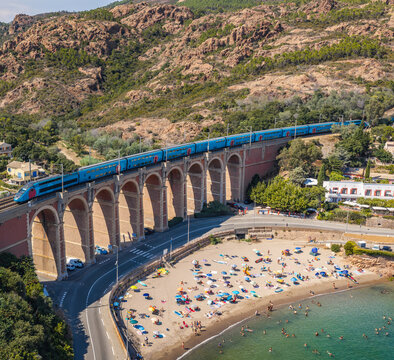 Aerial view of a high-speed train crossing a historic railway viaduct above a sandy beach with colorful umbrellas and turquoise water, Saint-Raphael, Provence-Alpes-Cote d'Azur, France.