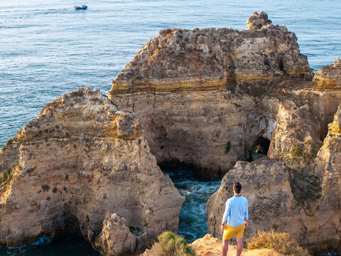 Aerial view of a man standing on a cliff edge overlooking dramatic limestone rock formations and the turquoise ocean in Faro District, Portugal.