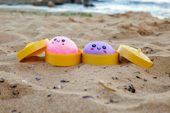 Pink and purple kawaii dumpling toys with cute smiling faces placed inside open yellow plastic capsules on a sandy beach outdoors
