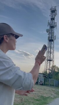 A focused woman in a cap uses her mobile phone outdoors, with a prominent communication antenna tower in the background under a vast, cloudy sky. She is connected to the digital world via wireless tec