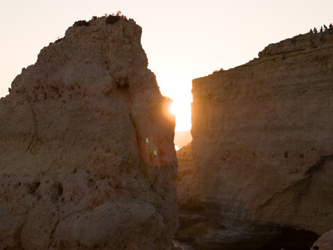 Aerial view of dramatic limestone cliffs at sunset with sun rays shining through a gap in the rock formation and people standing on the cliff edge in Faro District, Portugal.