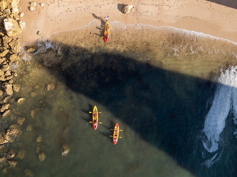 Aerial view of three colorful kayaks paddling in clear water near a sandy beach with rocky cliffs in Faro District, Portugal.