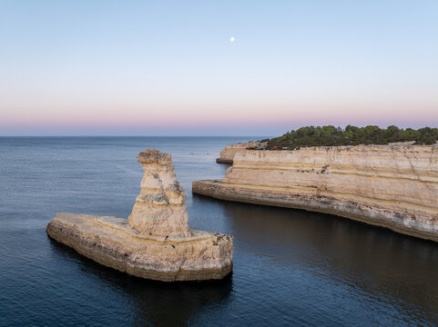 Aerial view of limestone sea stacks and cliffs rising from the calm ocean with sparse vegetation under a clear sky in Faro District, Portugal.