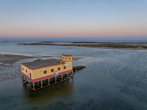 Aerial view of a yellow stilt house situated in the calm waters of the Ria Formosa lagoon, surrounded by sandy banks under a clear sky in Faro District, Portugal.