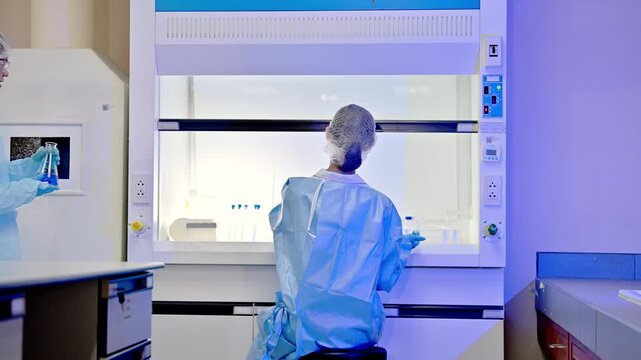 Scientists in sterile lab coats and hairnets work inside a fume hood with equipment