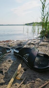 Black rubber sandals rest on wet beach sand near water showing textured waterproof soles. Concept of summer footwear, durable beach gear, outdoor relaxation, and vacation.