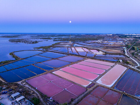 Aerial view of salt pans with vibrant pink and blue water pools under a twilight sky with a visible moon in Faro District, Portugal.