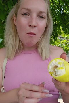 A woman with blonde hair displays a partially bitten lemon-shaped molded shell pastry; her puckered expression highlights the tangy flavor profile, framed by verdant greenery