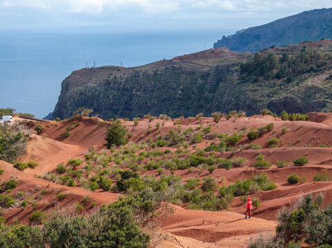 Aerial view of the volcanic landscape of La Palma with red soil, sparse shrubs, and a person in a red jacket standing on the terrain overlooking the ocean in Canary Islands, Spain.