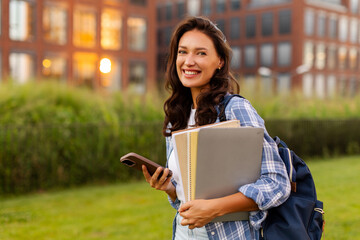 Fototapeta premium Cheerful young female student standing outdoors on campus holding smartphone and laptop