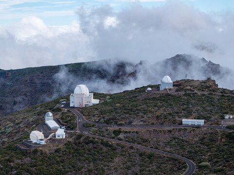 Aerial view of the Roque de los Muchachos Observatory featuring multiple telescope domes situated on a volcanic mountain ridge surrounded by low-hanging clouds in the Canary Islands, Spain.