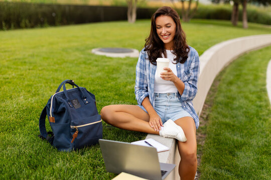 Smiling young female student relaxing with takeaway coffee while using laptop outdoors