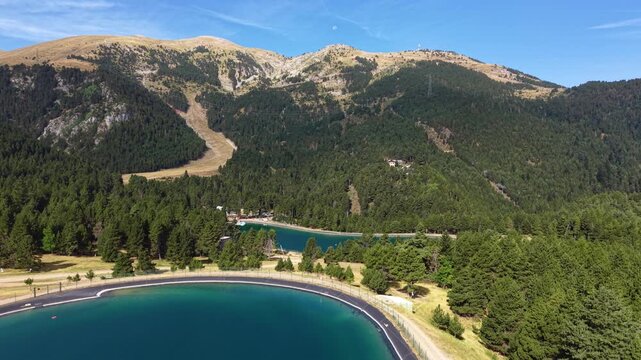 Panoramic view of La Molina ski resort in summer, Catalonia, Spain, featuring a turquoise mountain lake surrounded by pine forests and Pyrenees peaks under blue sky