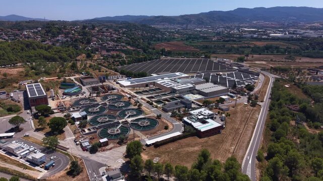 Overhead view of a large wastewater treatment facility featuring multiple circular clarifier tanks, solar panels on rooftops, and surrounding Mediterranean landscape with hills and town