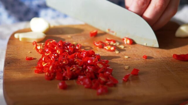 Hot red chili rings and garlic cloves being sliced on rustic wooden board in natural sunlight