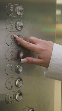 Hand pressing elevator buttons with braille and numbers. Brushed metal finish highlights tactile button layout. Lighting emphasizes metallic sheen under direct illumination