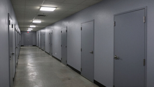 Empty prison corridor with heavy gray cell doors beneath stark fluorescent ceiling lights