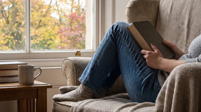 Woman reading a book while relaxing in an armchair beside a warm drink and autumn window. Cozy home leisure, comfort, mindfulness, and quiet seasonal lifestyle concept.