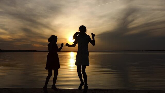 Two little sisters jumping happily in front of the setting sun and the big lake. Beautiful sunset.