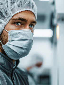 Zbliżenie na twarz mężczyzny z niebieskimi oczami, w białej maseczce ochronnej i czepku medycznym, w jasnym, sterylnym otoczeniu produkcyjnym lub laboratoryjnym.