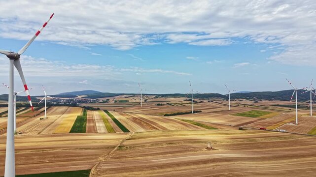 Wind turbines are turning in the open fields surrounded by crops on a sunny day. The landscape shows patches of brown and green land. Some mountains are visible in the distance.