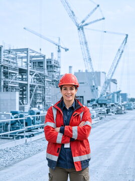 Smiling female construction worker with crossed arms at an industrial site. Vertical portrait of a woman engineer wearing a red hard hat and safety jacket near building framework and cranes