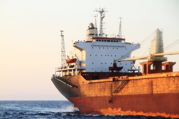 Large commercial cargo ship with rusted orange hull and white superstructure sailing on open blue sea with cranes visible © bulentbaris
