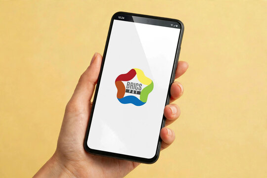 Ilustra&ccedil;&atilde;o de uma m&atilde;o feminina segurando smartphone exibindo o logo do BRICS Pay. Brasil, 19 de agosto de 2026.