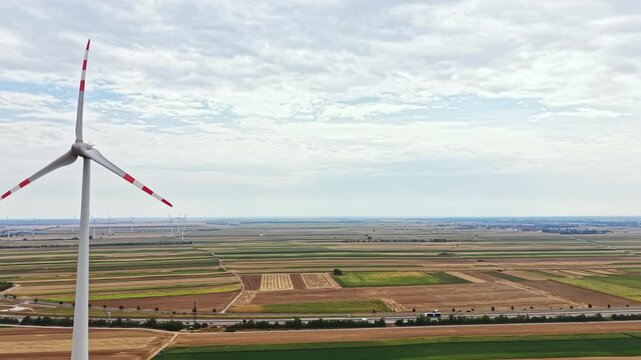 A wind turbine rotates slowly in a large field filled with various crops. Farmland extends into the distance, marked by lines and shapes. Clouds cover the sky with a hint of blue.