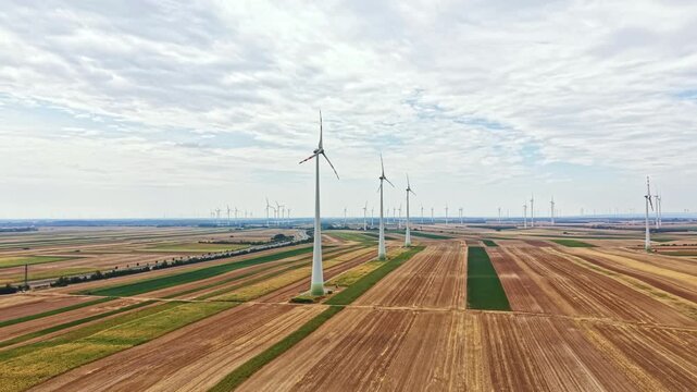 Wind turbines create energy in a flat farming area with rows of crops under a cloudy sky. The scene shows the large structures spaced apart across the land.