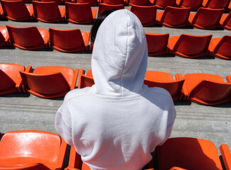 Fototapeta premium Person in white hoodie sitting in empty orange stadium seats outdoors