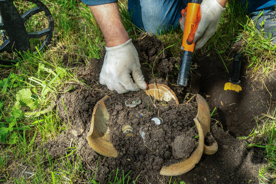 A treasure hunter is digging in the ground with a metal detector in a field. They are uncovering a piece of pottery filled with coins. The sun is shining and the ground is moist