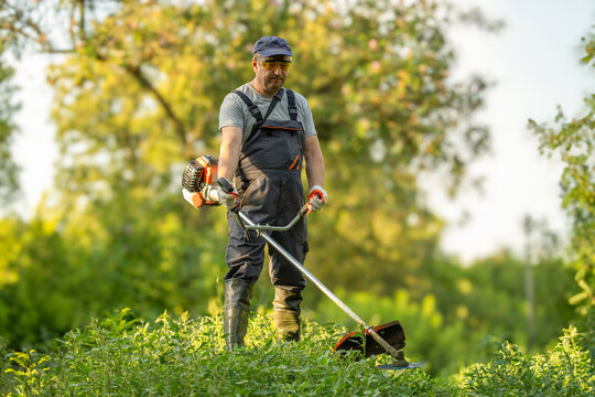 A working man stands on a hill using a brush cutter to trim grass in a field. The activity takes place during the day in a lush, green area surrounded by trees