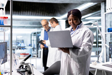 Fototapeta premium Black female researcher in a clinical research lab studies experiment data with a laptop, showcasing biotechnology, genetics research and pharmacology for progress in healthcare and medical research.