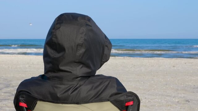 Person wearing hooded jacket relaxing on beach. Rear view of a solitary person in a black hooded jacket sitting on a folding chair at the beach, peacefully watching the calm sea waves under a blue sky