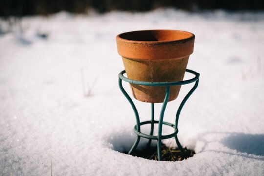 Empty terracotta pot rests on metal stand. Snowy ground surrounds garden planter. Winter scene hints at dormancy before spring growth begins.