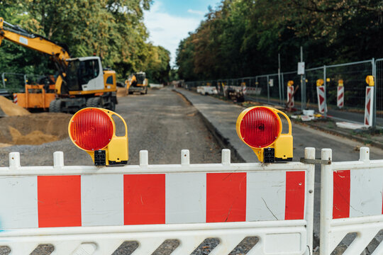 Red and white barrier with two yellow warning lamps closing off a road construction site with excavators, urban infrastructure renewal and traffic detour