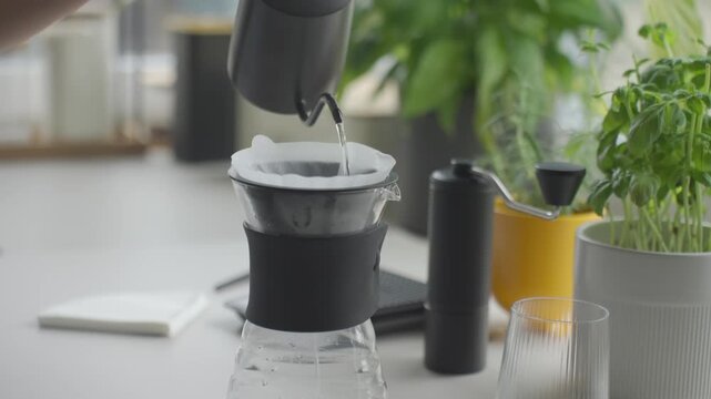 Hand pours water from kettle into paper coffee filter inside glass dripper on kitchen counter. Concept of pour over coffee preparation and filter rinsing