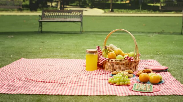 Empty city park contains a blanket and picnic basket on grass, with fresh fruit, pastries and juice arranged on a checkered blanket in the sunny urban park.