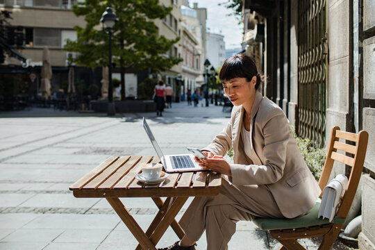 Professional woman working on laptop and phone at sidewalk cafe in city