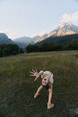 Naklejka premium Energetic woman bends toward the camera in a sunlit meadow, mountains in the background, casual outdoor wear, capturing a playful and adventurous lifestyle moment