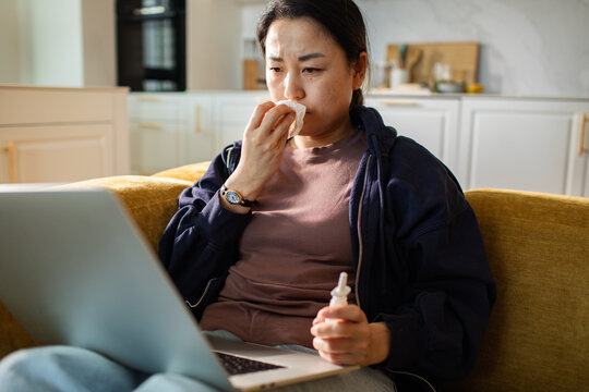 Woman using nasal spray and tissue while on laptop at home