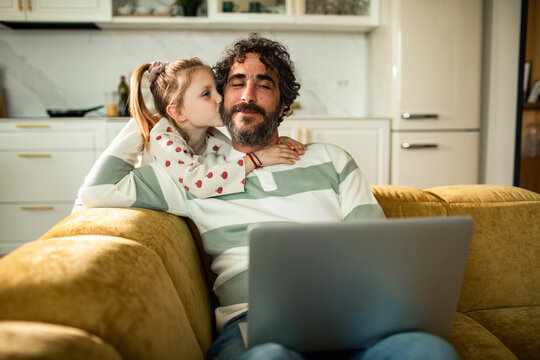 Father working on laptop at home while daughter kisses his cheek